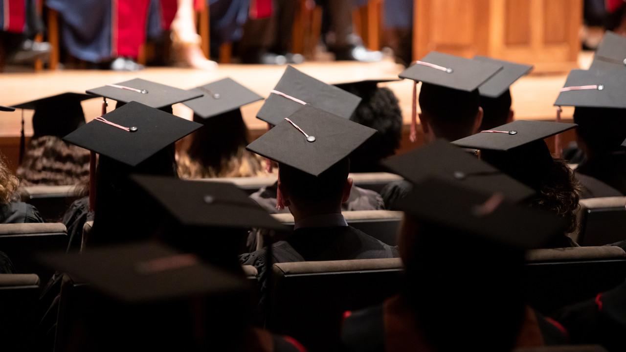 A group of graduates during a commencement ceremony. They are wearing traditional black caps and gowns. The setting is Alice Tully Hall with rows of seating filled with the graduates. The angle of the photo suggests it’s taken from the back of the hall, focusing on the back of the graduates' heads and their mortarboards, with one in the foreground serving as a focal point.