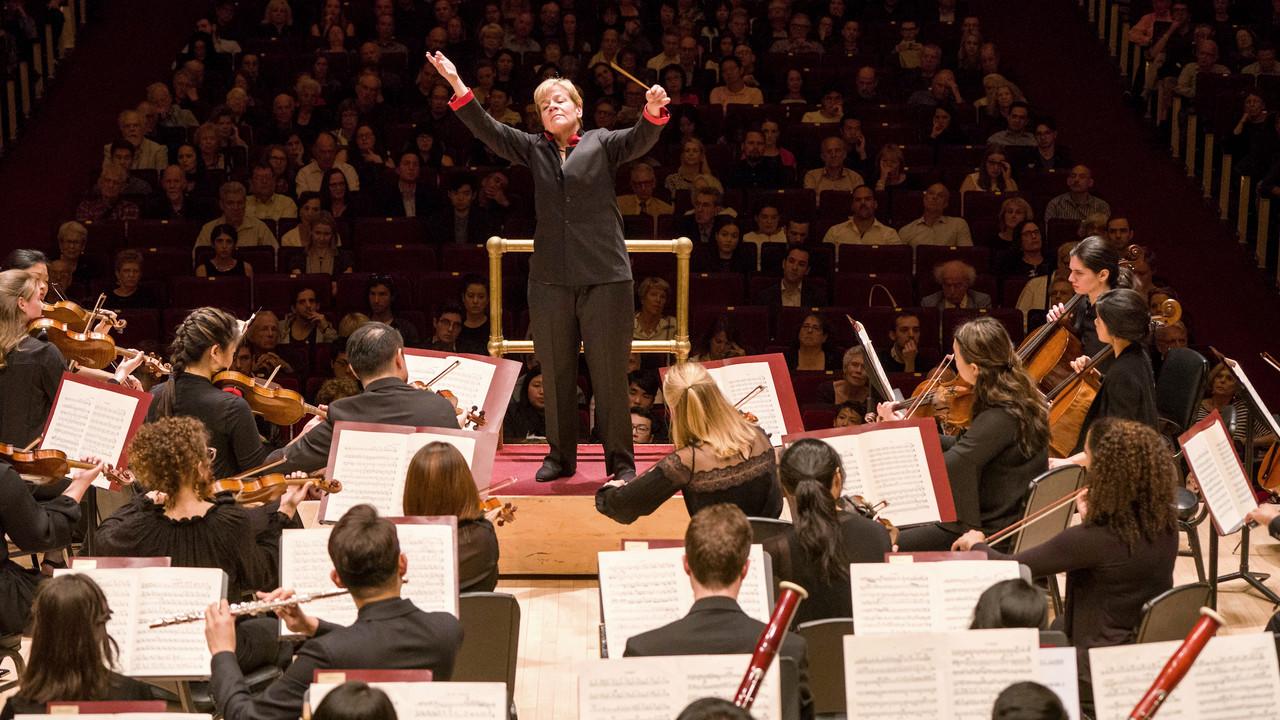 Juilliard Orchestra in the midst of a performance, with 3月in Alsop energetically leading the ensemble. 售票员, 位于中心位置, is dressed in a dark suit with their arms raised, 手里拿着指挥棒, 表达激情和命令. 音乐家, focused 和 playing their instruments, are arrayed in a traditional orchestral setup on the stage of a concert hall. 观众挤满了剧院, witnessing the event in a well-lit 和 gr和 venue, which adds to the formal 和 prestigious atmosphere