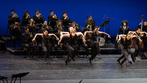 A vibrant performance featuring a group of 舞者 in black, 亮片服装, energetically executing a dance routine on stage. 在他们身后, a big band with brass instruments provides live music, creating a dynamic backdrop for the 舞者. The setting suggests a collaborative concert or musical performance, highlighting the intersection of live music and dance.