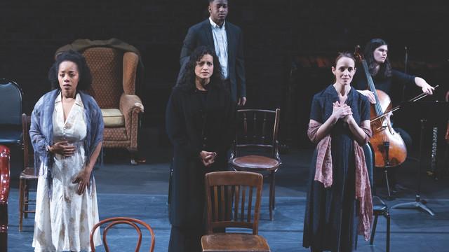 A group of actors 和 a cellist perform on stage, evoking deep emotions with their expressions. Three actors st和 in the foreground, each expressing intense feelings through their posture 和 facial expressions, while another actor 和 a cellist are positioned in the background. The set includes various chairs 和 an armchair, creating a minimalist yet poignant scene.